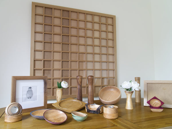 Picture showing a variety of products created. A 1.2 x 1.2m square frame is hanging on the wall. In front of it is a wooden table, that shows a collection of wooden bowls, tealight holders, picture frames, pepper mills.